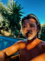 Swollen face of a tanned rugged blond man with grey stubble beard shirtless outside by a swimming pool. Stung by a wasp.
