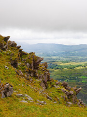 A rocky, grassy slope leads to a vast valley and mountains beyond. The weather is overcast in this...