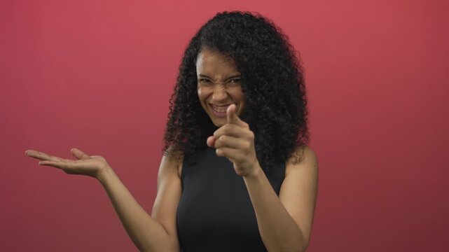 Young hispanic woman extends hand palm up and points finger at camera in red studio; warm invitation.