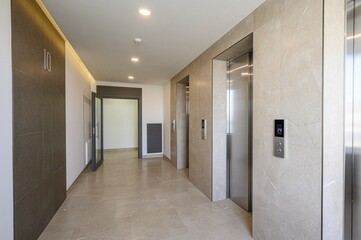 modern hallway with beige marble walls and floors features elevator doors on the right and an open doorway on the left, revealing a bright interior
