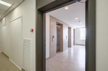 view through a doorway shows a modern hallway with elevators. The space is well-lit with tile...
