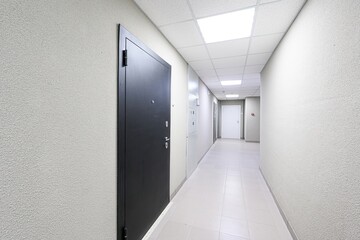 well-lit hallway with a dark door on the left. The walls are textured, and the floor is tiled. Ceiling panels with lights run the length of the hallway, leading to a white door in the distance