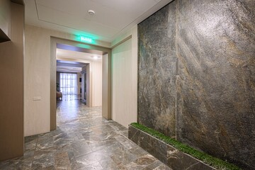 modern hallway features stone flooring, a textured wall with plants, and an exit sign in green. An open doorway leads to a bright living area