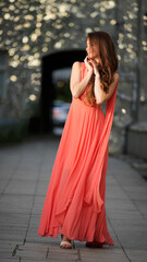 Graceful young woman in coral gown enjoying a quiet moment on a city street with bokeh light...