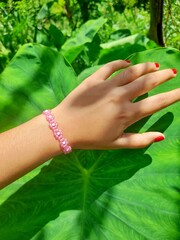 A hand wearing a pink flower bead bracelet rests on a large green leaf, creating a natural and...