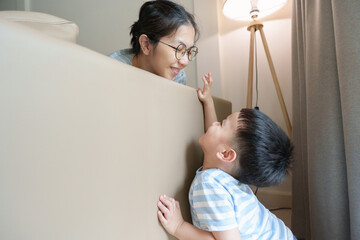 Laughing Asian mother and young boy bonding together behind sofa, playing peekaboo in living room.
