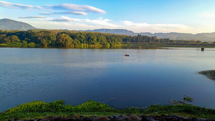 A truly beautiful morning view. The sky is so clear and blue. The reservoir is set against a backdrop of hills and mountains. Someone is fishing in a boat.