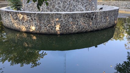 A circular stone wall, part of a water feature or fountain, reflects on the still water below, with...