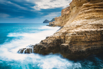The ocean is calm and the sky is cloudy. Waves breaking on the cliff