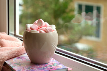 Marshmallows in a mug on a balcony windowsill. A mug with marshmallows in the morning against the background of house windows. Dessert close-up.