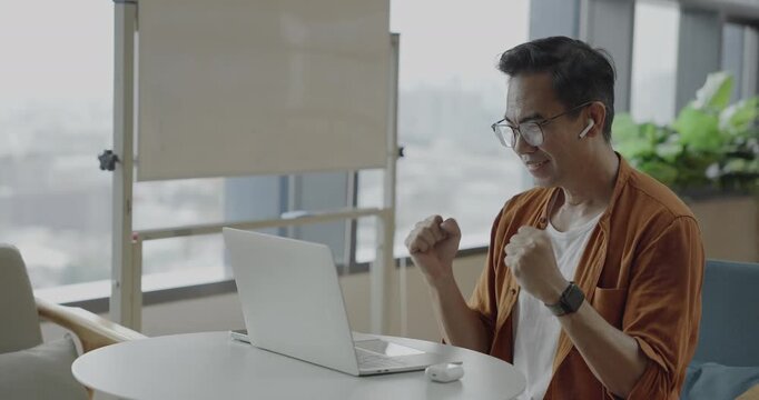 Senior Asian businessman wearing headset celebrates success during online meeting in modern co-working space. Remote work and business communication concept.