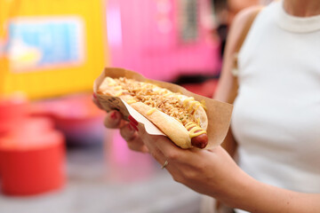 Close-up of woman holding delicious hot dog with toppings in brown paper tray at lively carnival or...