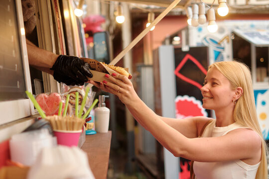 Joyful young blonde woman exchanges cash for food at lively outdoor market food truck with string stringlights, warm glow creating friendly, festive atmosphere