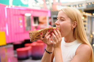 Young woman with long blonde hair biting hot dog with toppings in colorful outdoor market with...