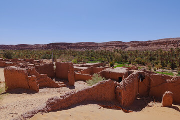 Ancient Mud Brick Ruins in Algerian Desert Oasis with Palm Trees and Rocky Cliffs Under Clear Blue...