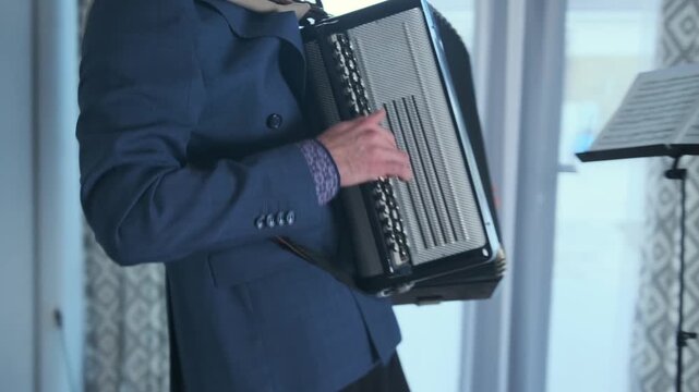 Close-up of male hands playing the bayan accordion while standing in a modern living room. The musician in a suit performs expressive classical music in front of a music stand with sheet music
