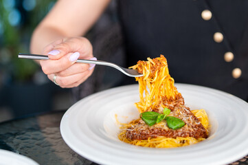 Close-up of spaghetti with meat sauce and basil in a white plate, held by a fork, with a blurred...