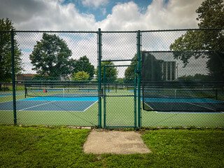 Gate leading to empty pickleball courts. Focus on the fence and door leading to the new courts. Public park setting in Summer with cloudy blue skies.