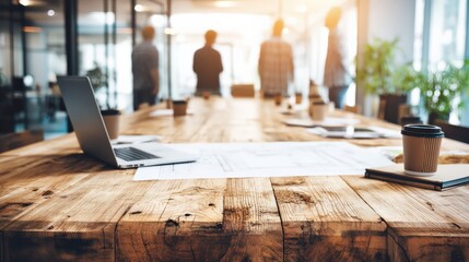 Modern office meeting room with a wooden table, laptop, coffee cup, and blurred group discussing in the background with natural sunlight.