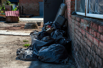 Black trash bags are stacked against a weathered brick wall, showing neglect in an empty industrial space under bright sunlight