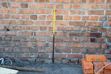 A yellow-handled tool stands upright near a brick wall, surrounded by construction materials and a dust-covered ground