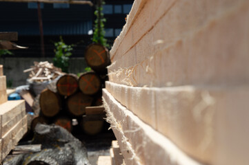 Sturdy wooden planks are neatly stacked at a lumber yard, showcasing fresh cuts and surrounded by round logs under bright sunlight