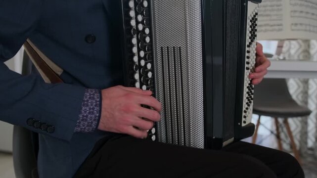 Close-up of a bearded man in glasses playing the bayan accordion at home. He sits in the kitchen, swaying to the rhythm, with emotional focus on the music. Sheet music is visible in the background.