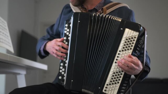 Bearded young man studies music at home, practicing on a accordion, sits in a modern kitchen with a music stand in front of him, focusing on technique and rhythm. Home learning and music education.