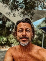 Smiling tanned man with stubble beard and blond hair enjoying summer garden with olive trees.