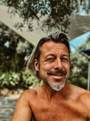 Smiling tanned man with stubble beard and blond hair enjoying summer garden with olive trees.