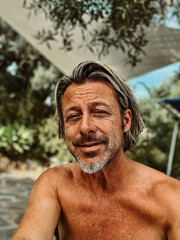 Smiling tanned man with stubble beard and blond hair enjoying summer garden with olive trees.
