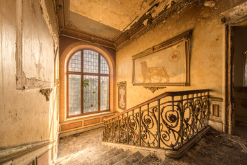 Sunlit abandoned staircase with ornate wrought iron railing and large arched window in historic...