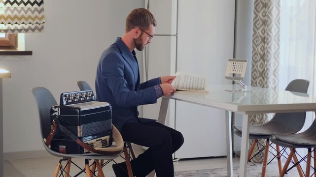 Stylish bearded man in a suit reads and flips through sheet music while sitting at a modern table. An accordion rests on a chair nearby. Calm and intellectual atmosphere.
