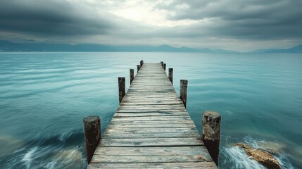 Fototapeta premium Old wooden pier extending into calm turquoise lake under dramatic cloudy sky with mountains in the distance