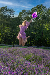 A person wearing a flowing purple dress jumps joyfully in a vibrant lavender field, holding a matching hat. The lavender blooms stretch out in the foreground, 