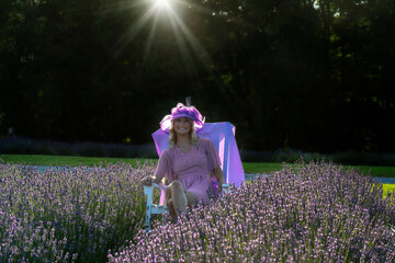 A woman in a pink dress and hat sits on a chair amidst a lush lavender field, with sun rays gently illuminating the scene from above. The surrounding greenery creates a peaceful and serene backdrop, 