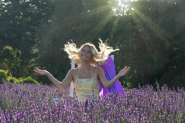A woman with blonde hair stands joyfully among vibrant lavender flowers, with sunlight streaming through the trees behind her. She wears a floral dress and appears relaxed and happy,