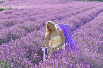 A woman in a light summer dress sits on a chair surrounded by endless rows of blooming lavender....