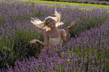 A smiling woman with flowing hair sits amidst a field of vibrant lavender flowers, creating a joyful and serene atmosphere. Sunlight enhances the scene with a warm, natural glow.