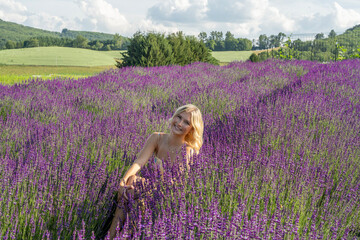 A woman with blonde hair is sitting amidst a vibrant lavender field, enjoying a sunny day. Rolling hills and a few scattered clouds can be seen in the background, enhancing the serene atmosphere.