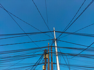 Low angle view of electricity poles and telecommunication poles filled with a tangled and disorderly network of cables, contrasting with the bright blue sky background.