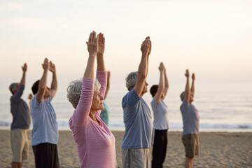 A group of older adults practicing yoga on the beach during sunrise or sunset, raising their arms in a peaceful, focused pose.