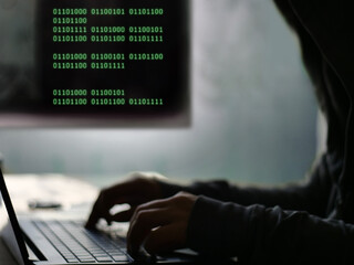 Cybersecurity Threat: Hacker's Hands on Keyboard with Encrypted Digital Information - Mysterious Hooded Figure Typing on a Laptop with Green Binary Code Overlay on Screen
