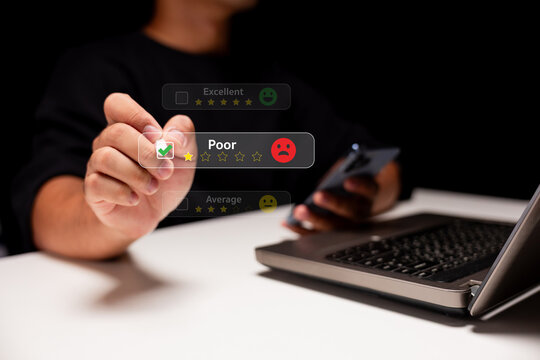A person uses a digital interface to rate a service as "Poor" with one star, while holding a smartphone near a laptop on a white desk. - Powered by Adobe