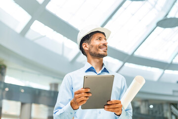 Young smiling man architect wearing a hard hat making a note in digital tablet with blueprint plans...