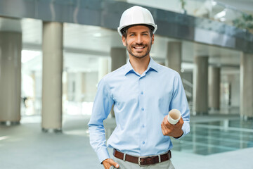 Close-up portrait of a young smiling man architect wearing a hard hat with blueprint plans looking...