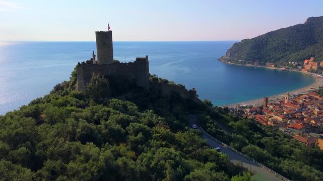 Aerial view of Monte Ursino castle and village on the Italian Riviera. Coastline with beach umbrellas, Mediterranean Sea. Green hills in the background. Vacation. Tourism. Noli Liguria Italy 1.09.2025