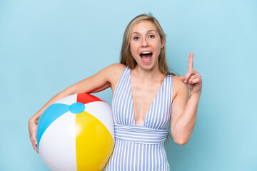 Young blonde woman in swimsuit holding beach ball isolated on blue background pointing up a great...
