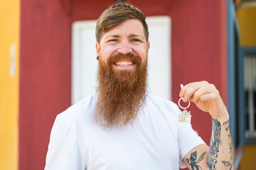 Redhead man with beard holding home keys at outdoors smiling a lot