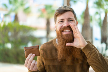 Redhead man with beard holding a wallet at outdoors shouting with mouth wide open